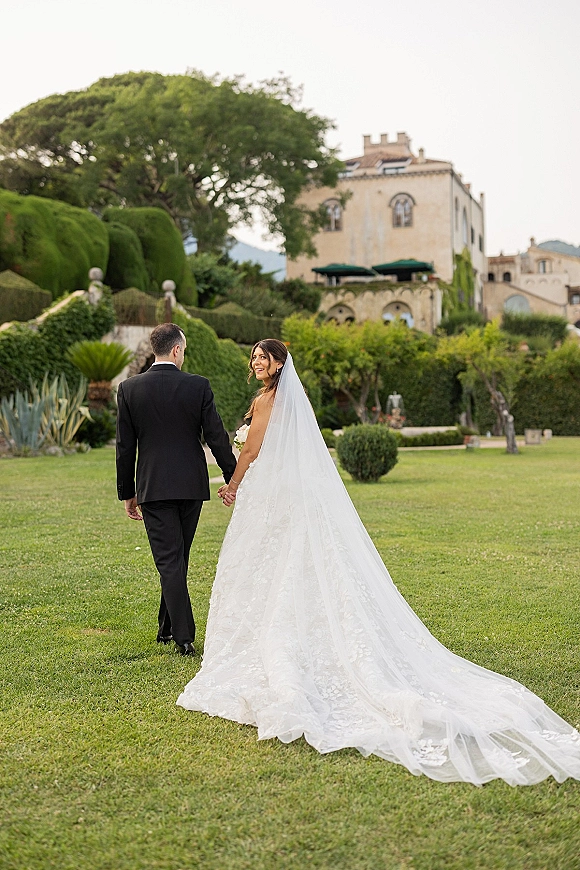 Couple portrait of newlyweds holding hands, walking on a lawn as the bride looks back with a long veil near a stone estate building