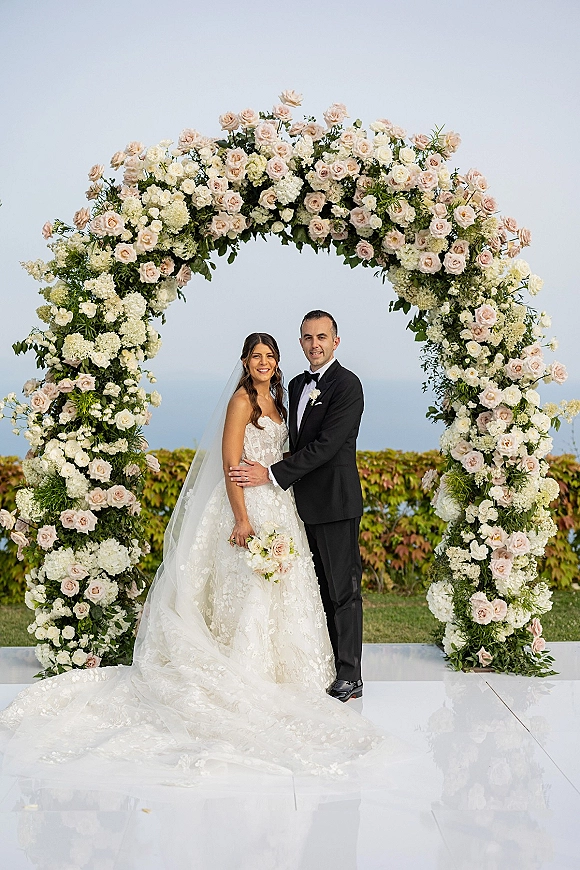 Couple portrait of bride and groom under arch, hugging beneath a round rose floral arch on a white platform with ocean behind