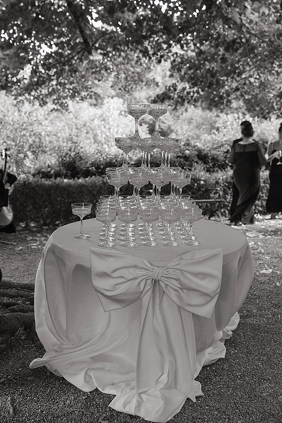 Champagne tower of stacked coupe glasses on a round table with a white tablecloth and fabric bow, set along a garden path with guests