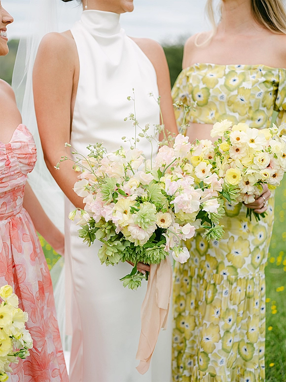 Bride with bridesmaids holding bouquets of pastel flowers and greenery, bride in high neck dress and veil in an outdoor field setting