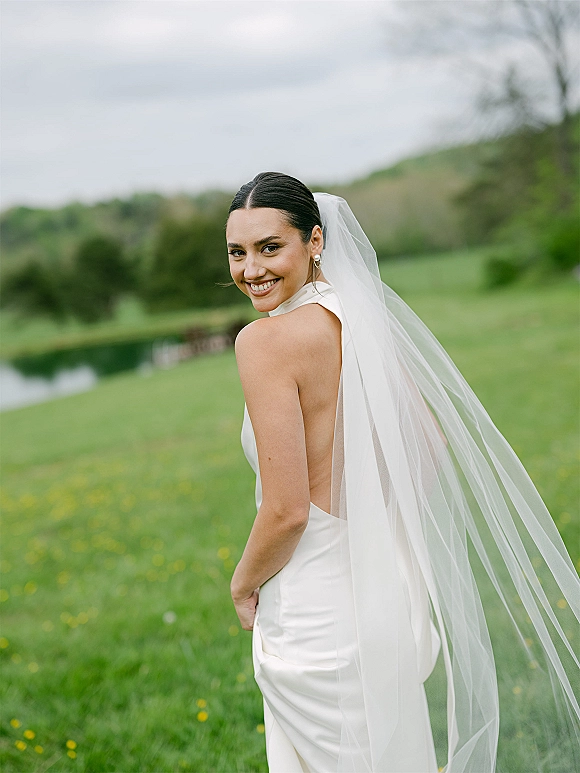 Bridal portrait of a bride looking back in a long veil, wearing pearl drop earrings, standing by a pond in a green meadow under clouds