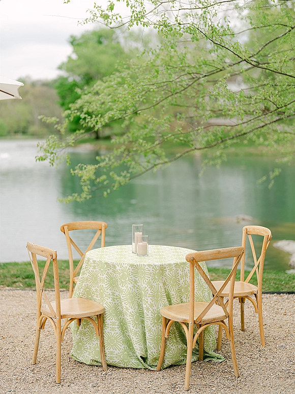 Outdoor reception table with a green patterned tablecloth, wood cross back chairs, and glass cylinder candles by the lake on a gravel patio