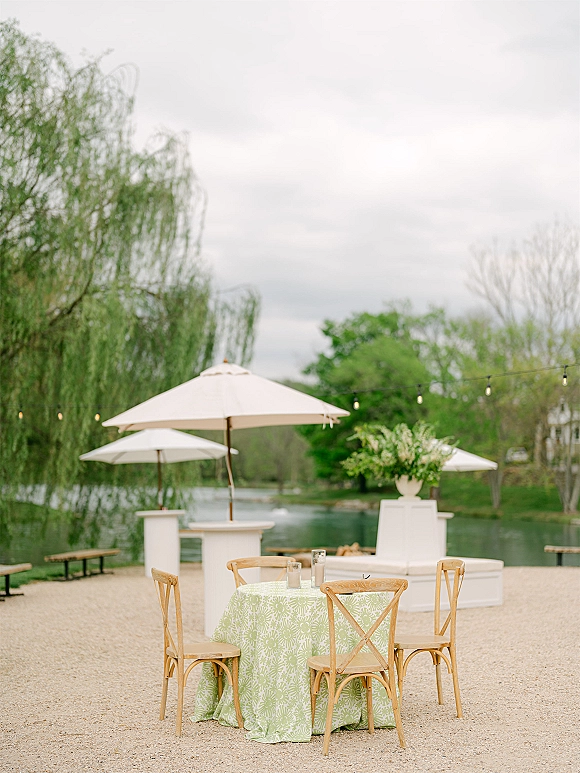 Outdoor reception setup with a round table in a green patterned tablecloth and cross back chairs under white umbrellas by the lake