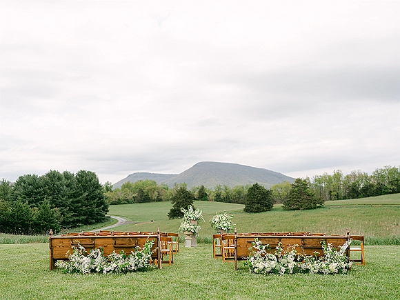 Outdoor ceremony setup with garden ceremony seating, wooden benches and chairs lining a floral aisle in a meadow with mountains under clouds