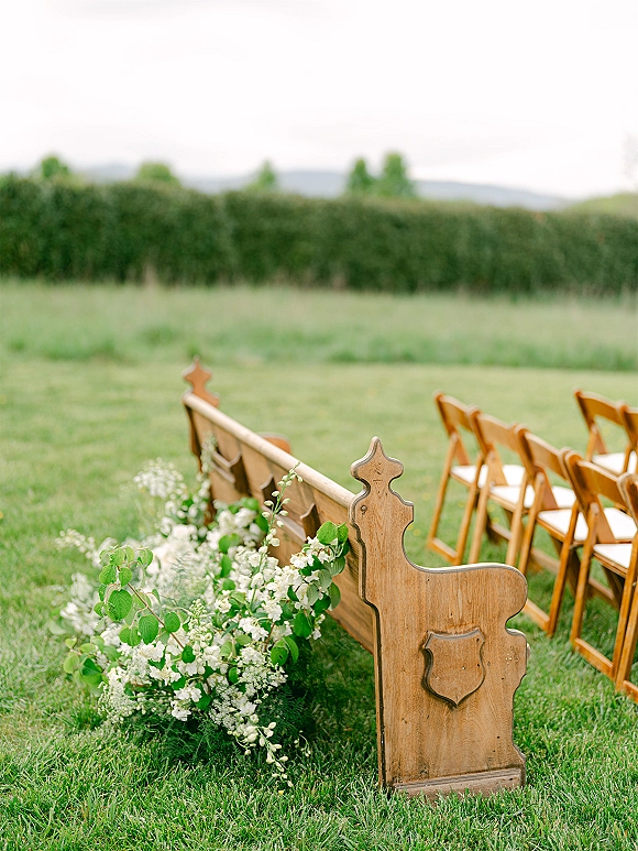 Ceremony seating with wooden pew and folding chairs beside white flower and greenery ground arrangements on a lawn with hedges and hills