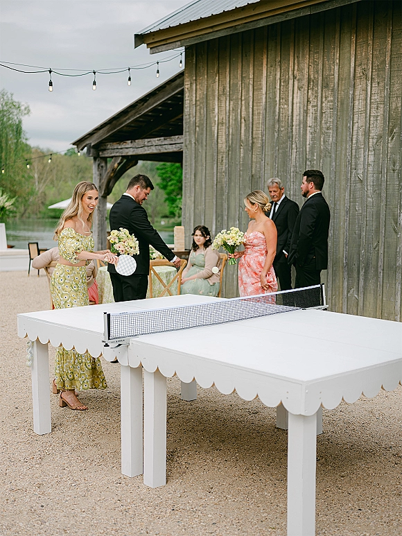 Wedding guest games as bridesmaids and men in black suits play wedding ping pong on a white table under string lights by a rustic barn wall