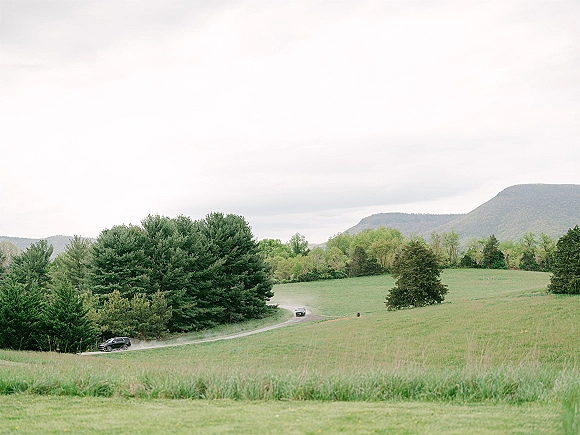 Countryside landscape with rolling hills and a winding road with cars through green fields, trees, and distant mountains under cloudy sky