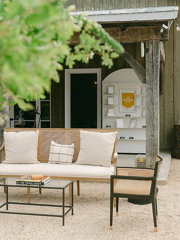 Wedding lounge seating with a cane sofa, throw pillows, and coffee table with books beside an escort card display under string lights by a barn doorway