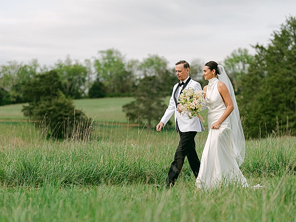 Wedding processional with bride walking with father, holding a bouquet and long veil, crossing a meadow field under a cloudy sky