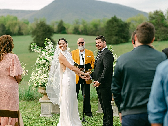 Wedding ceremony with bride and groom holding hands as officiant reads at microphone, framed by white floral urns and mountain view