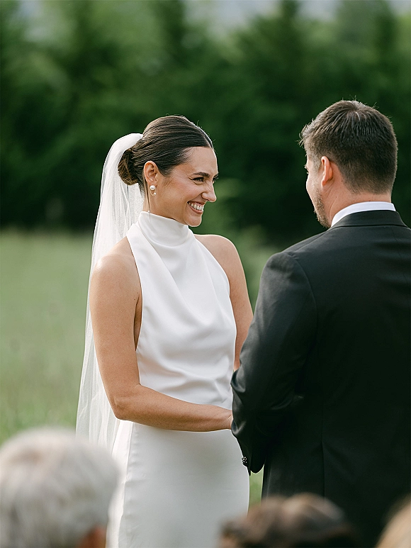 Wedding vows during an outdoor wedding ceremony as bride in veil and pearl earrings holds groom’s hand in a field with trees behind