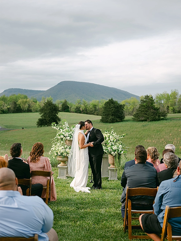 Ceremony kiss as bride in wedding dress and veil kisses groom in tuxedo at outdoor wedding ceremony with urn florals and mountain backdrop