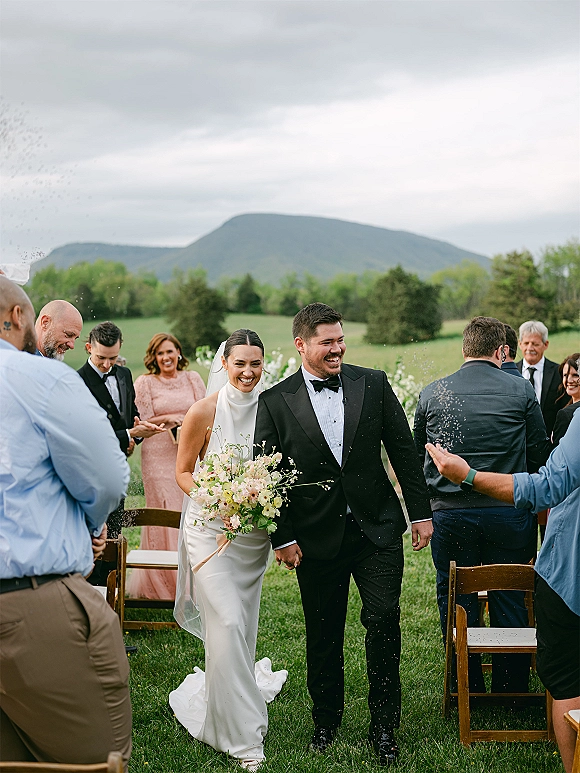 Wedding recessional as newlyweds walk down the aisle hand in hand, bride with bouquet and veil beside guests on a mountain lawn