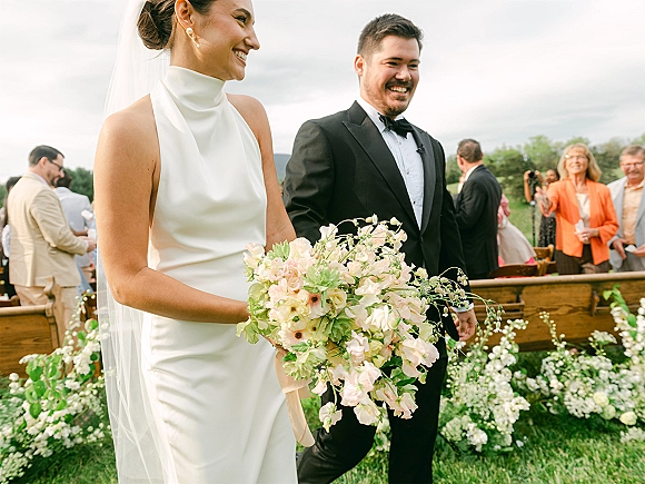 Wedding recessional as bride and groom walk the aisle, bride holding pastel bouquet and long veil, guests cheering on a cloudy lawn