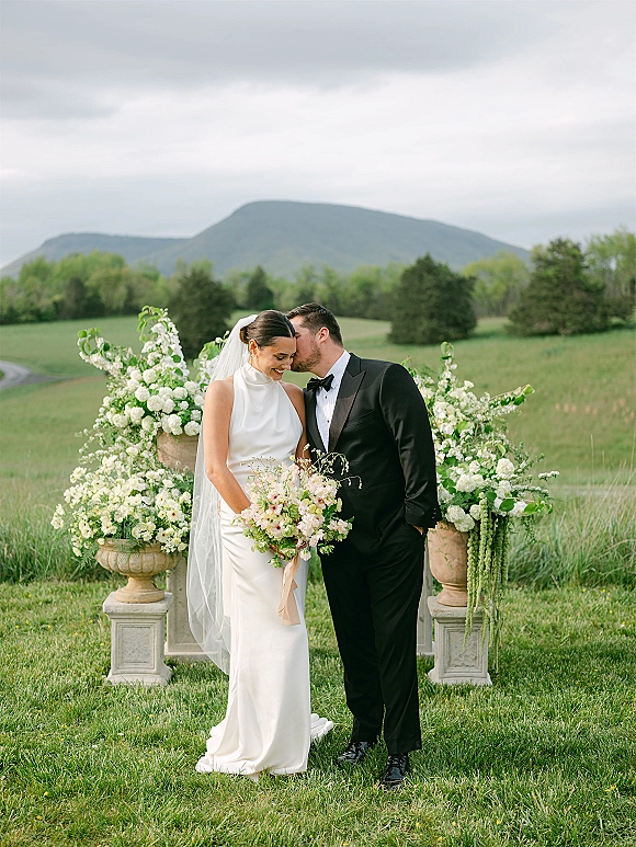 Couple portrait of groom kissing bride’s forehead as she holds a bouquet, veil flowing, with mountains and stone urn florals behind