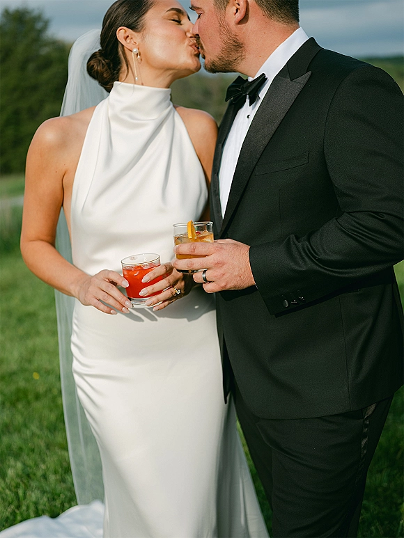 Wedding kiss portrait of bride and groom kissing while holding cocktails, her veil and pearl earrings glowing on a green lawn with hills