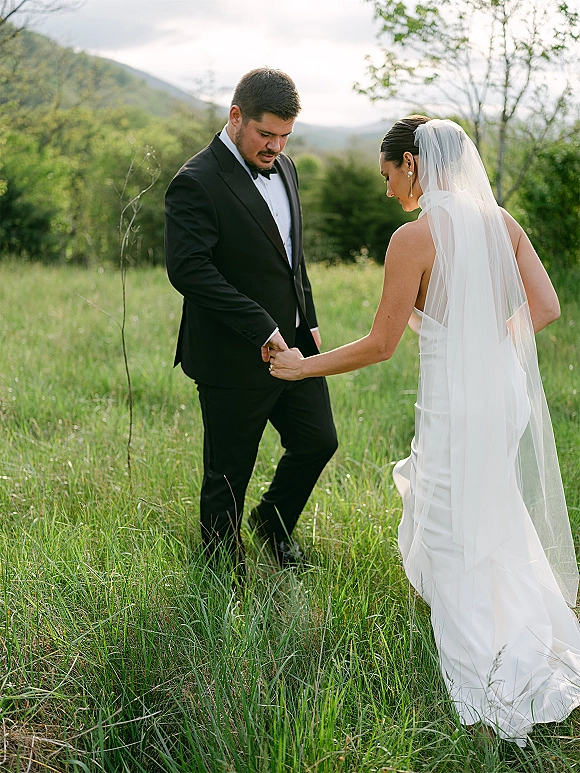Couple portrait of bride and groom holding hands, her long veil trailing behind, walking through a grassy meadow with mountains under cloudy sky