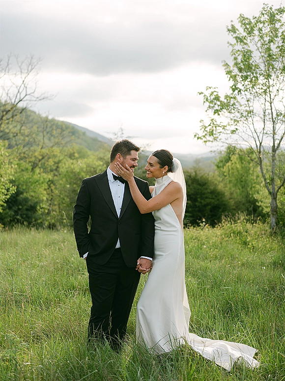Couple portrait of bride and groom holding hands, bride in veil touching his face in a meadow with mountains under cloudy sky