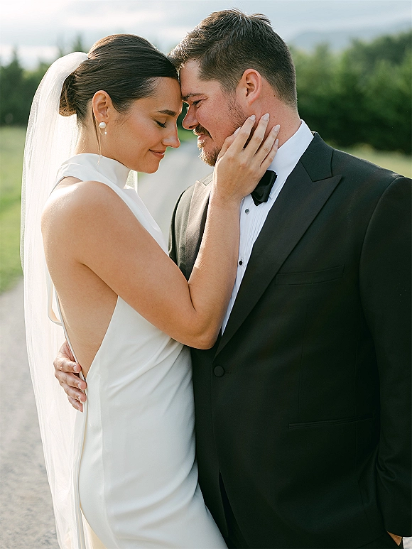 Couple portrait of bride and groom close up, forehead touching as she cups his face, veil and pearl earrings on a tree-lined road