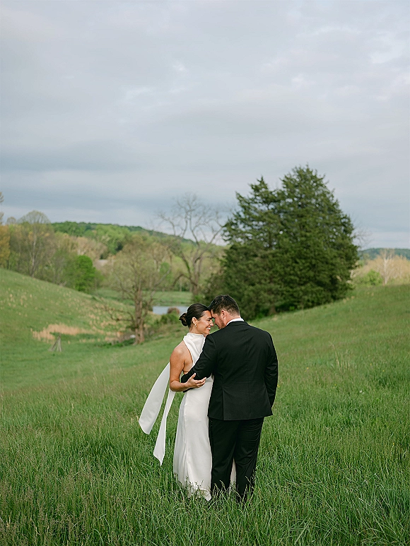 Couple portrait of bride and groom outdoors embracing in tall grass, bride in halter gown with scarf tie, rolling hills under cloudy sky