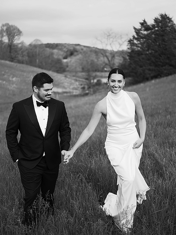 Couple portrait of bride and groom walking hand in hand, bride lifting her wedding dress in a meadow with rolling hills under cloudy sky