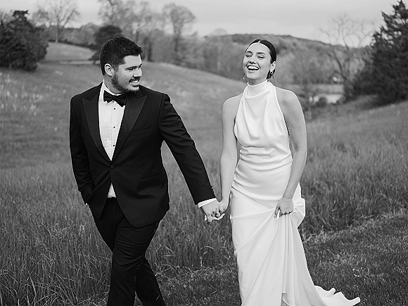 Couple portrait of bride and groom walking hand in hand, bride lifting her wedding dress beside him in a grassy field with rolling hills