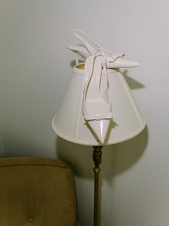 Bridal shoes, white wedding heels with pointed toes and bow detail perched on a table lamp beside a chair against a wall
