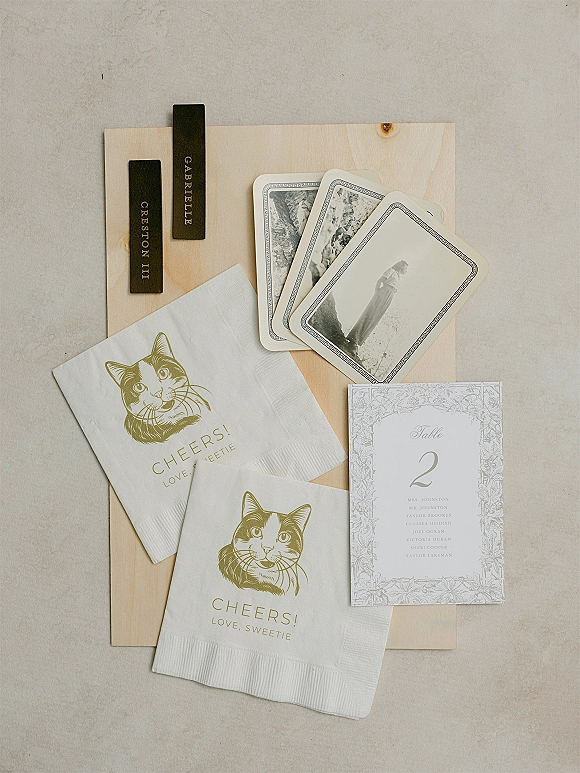 Wedding stationery flatlay with gold foil napkins, cat illustration, place cards and black tags arranged on a linen fabric surface
