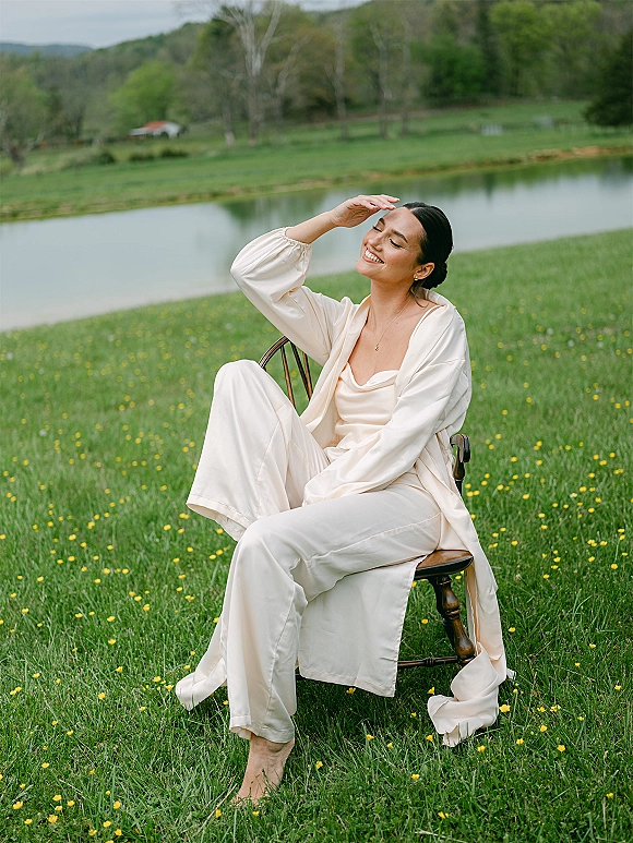 Bridal portrait of a bride in an ivory satin pajama set with a gold necklace, seated on a wooden chair in a wildflower meadow by a pond