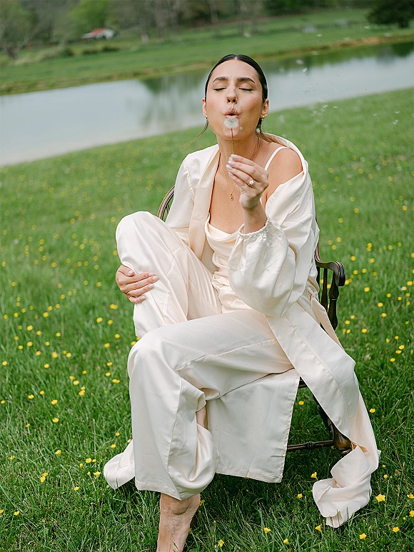 Bridal portrait of a bride blowing dandelion in a satin slip dress, seated on a wooden chair in a meadow by a pond