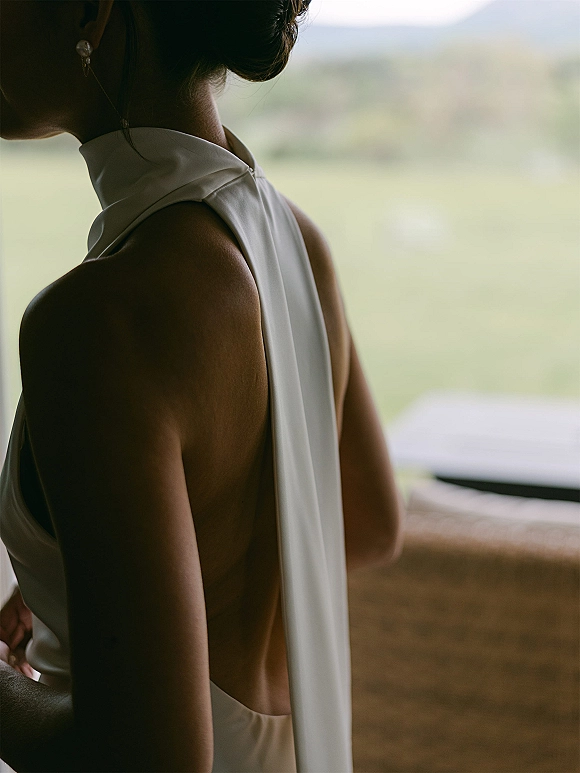 Wedding dress detail on a bride in a backless wedding dress with high neck collar, draped sash, pearl earring, in window light