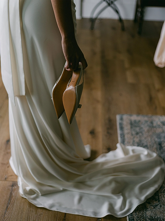 Bridal shoes, wedding heels in white pointed toe style resting beside a wedding dress train on a wood floor near an area rug