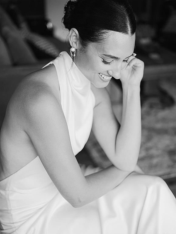 Bridal portrait in black and white of a bride laughing in a high-neck wedding dress with pearl drop earrings, seated on a bed in a bedroom