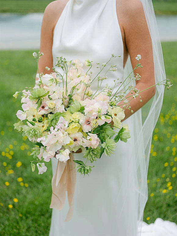 Bridal bouquet of pastel wedding bouquet blooms with greenery and a long ribbon, held against a white dress and veil by water and wildflowers