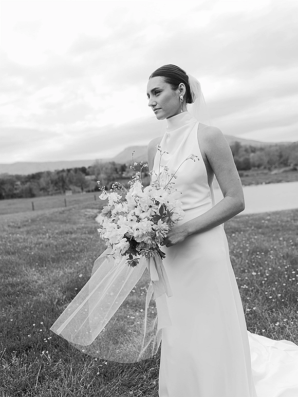 Bridal portrait of a bride holding bouquet with long ribbon, veil flowing as she looks away by a lakeside meadow and mountains under clouds