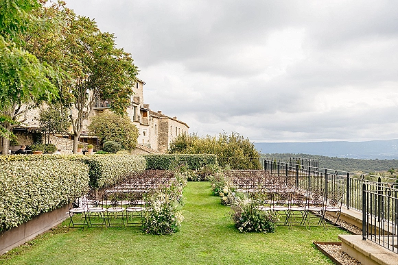 Ceremony setup for an outdoor wedding ceremony with wrought iron chairs and a floral-lined aisle beside hedges at a stone villa overlook