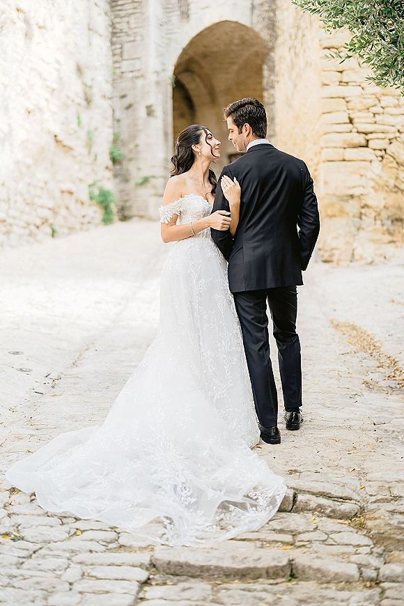 Couple portrait of bride and groom walking away under a stone archway, bride holding his arm, lace dress train trailing on cobblestones