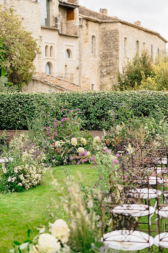 Outdoor ceremony setup with garden wedding ceremony chairs and meadow-style floral arrangements on grass beside hedges near a stone villa