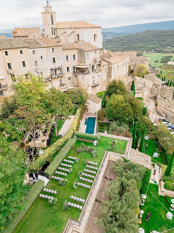 Outdoor ceremony setup with white folding chairs and aisle floral arrangements on a garden lawn by a pool, stone buildings, and valley hills beyond