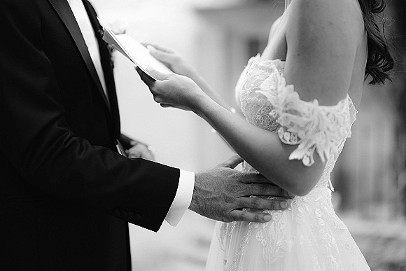 Wedding vows as bride reading vows from a vow book close up, groom in black tuxedo listening, lace off-shoulder gown, outdoor blurred architecture