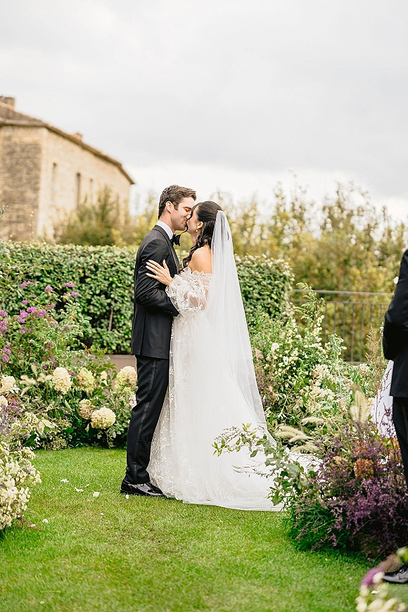 Wedding kiss portrait of bride and groom kissing, her cathedral veil and lace dress against hedges, lawn, and stone estate backdrop