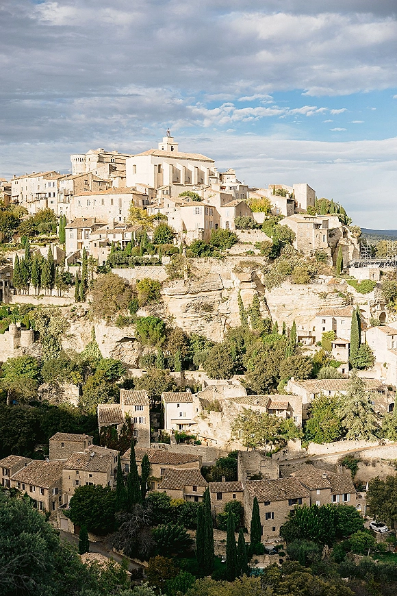 Hillside village view with a stone village skyline, terracotta rooftops and a church tower above cypress trees on a rocky cliff under blue clouds