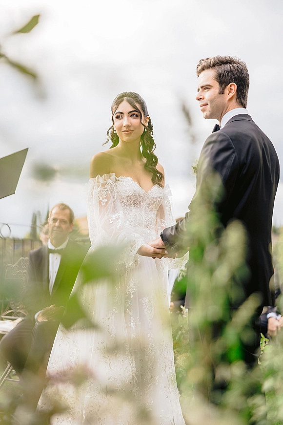 Ceremony moment at an outdoor wedding ceremony as the bride and groom hold hands, her lace off-shoulder dress and veil with guests seated behind.