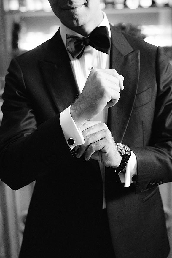 Groom getting ready, adjusting cufflinks on a black tuxedo with bow tie and wristwatch in soft window light, indoor room blurred behind