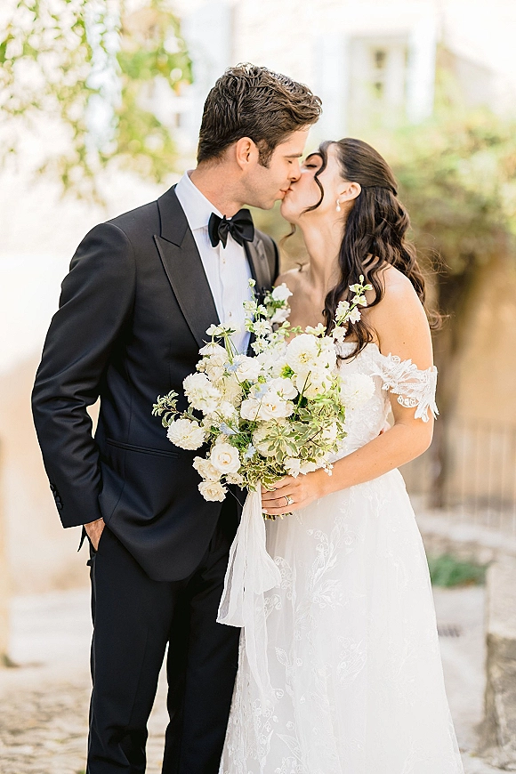 Wedding kiss portrait of bride and groom kiss, her lace off-shoulder gown and bouquet with pearl earrings on a stone walkway with greenery