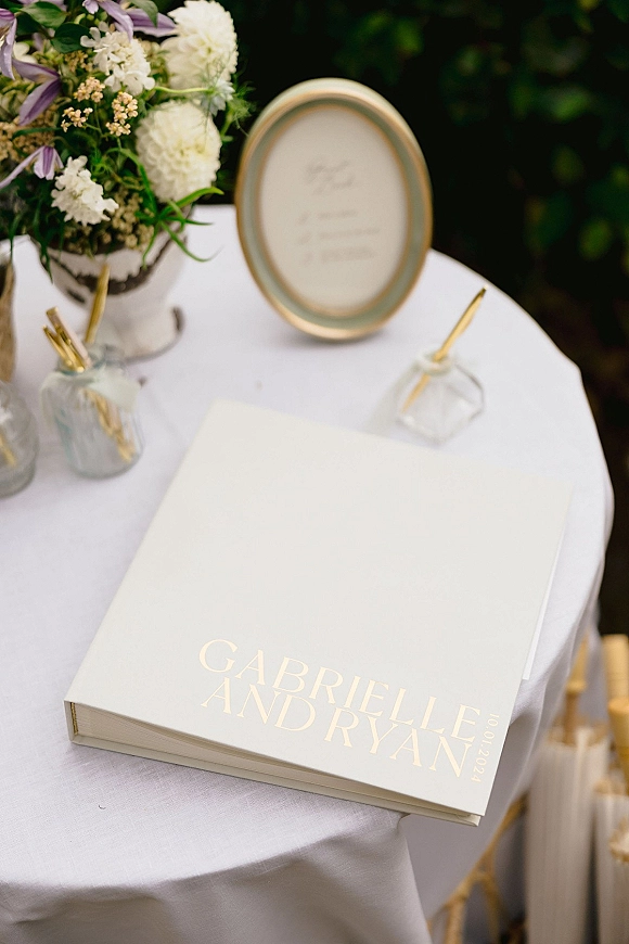Wedding guest book with gold foil lettering on a white linen welcome table, accented by white flowers, greenery, and a gold-framed sign outdoors