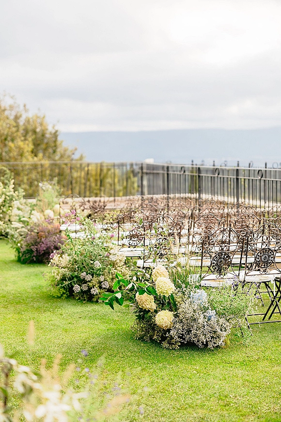 Ceremony aisle decor with wrought iron chairs and hydrangea floral arrangements lining a lawn aisle, set against shrubs and distant hills
