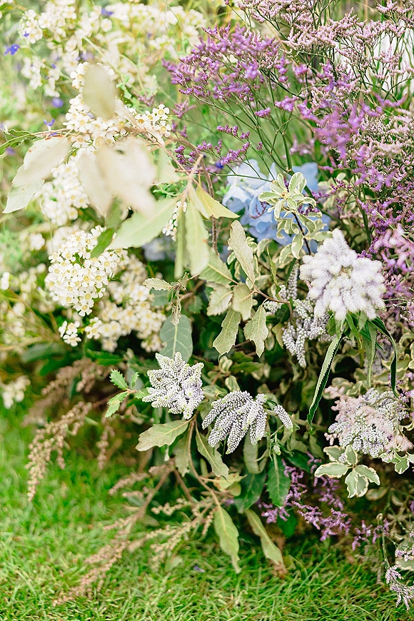 Wedding floral arrangement with ceremony ground florals featuring blue hydrangea, purple and white blooms, and greenery on a garden lawn