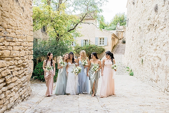Bride with bridesmaids walking together, holding white and green bouquets with ribbon streamers along a cobblestone street by stone walls