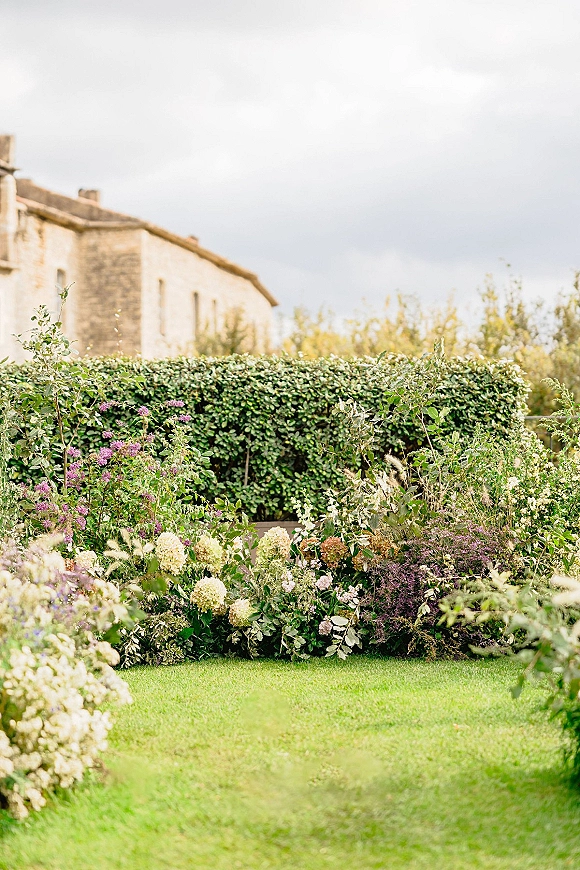 Garden ceremony aisle with wildflower and hydrangea arrangements lining the lawn, framed by manicured hedges near a stone building under cloudy sky